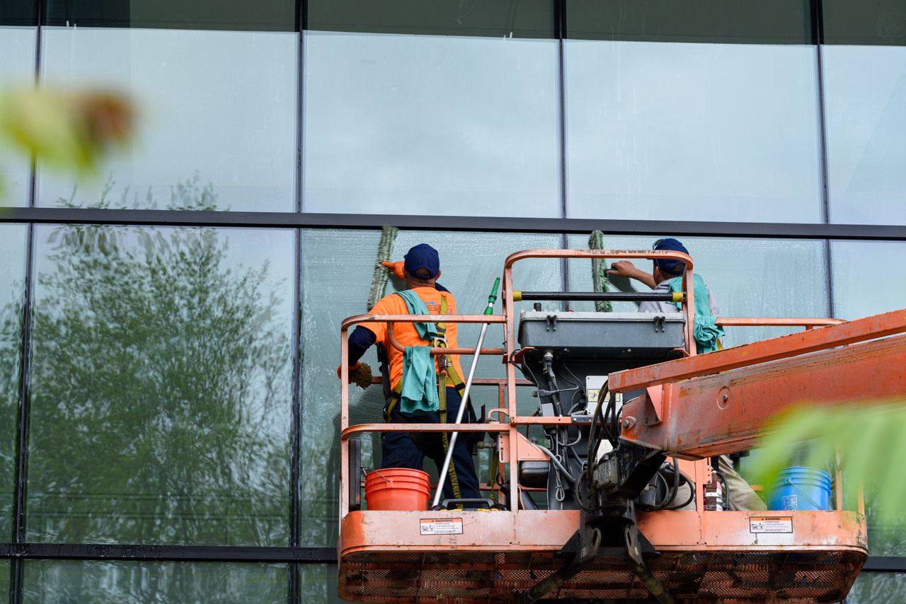 brand-01 Two workers cleaning windows on a glass building facade using a lift platform, enhancing building aesthetics.